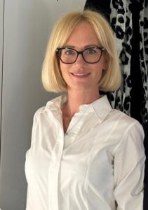 Frau mit blonden Haaren, Brille und weißer Bluse lächelt freundlich.